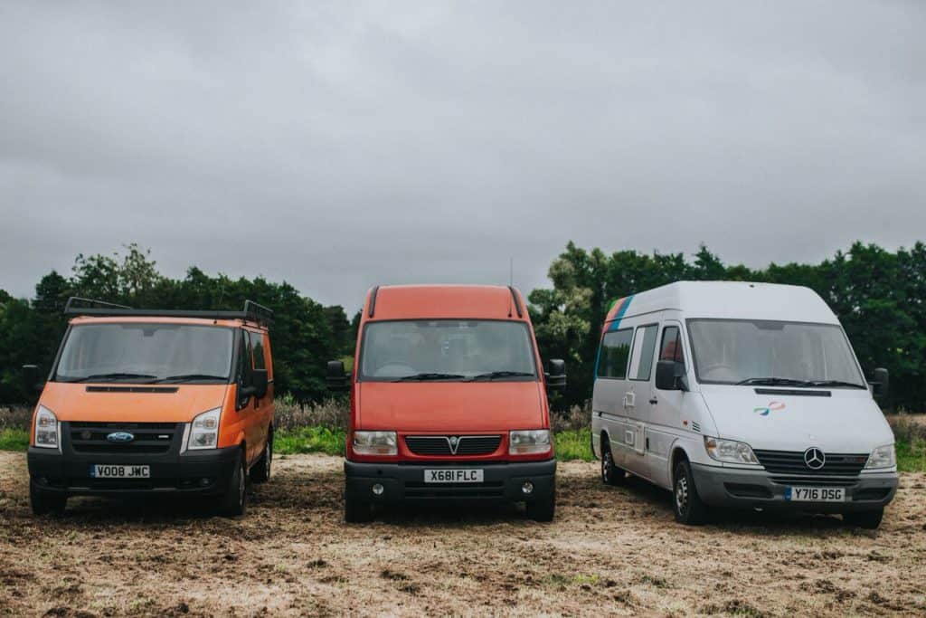 Three vans on a campervan trip parked in a grassy area. The vans are orange, red, and white, from left to right. Trees and an overcast sky are visible in the background.