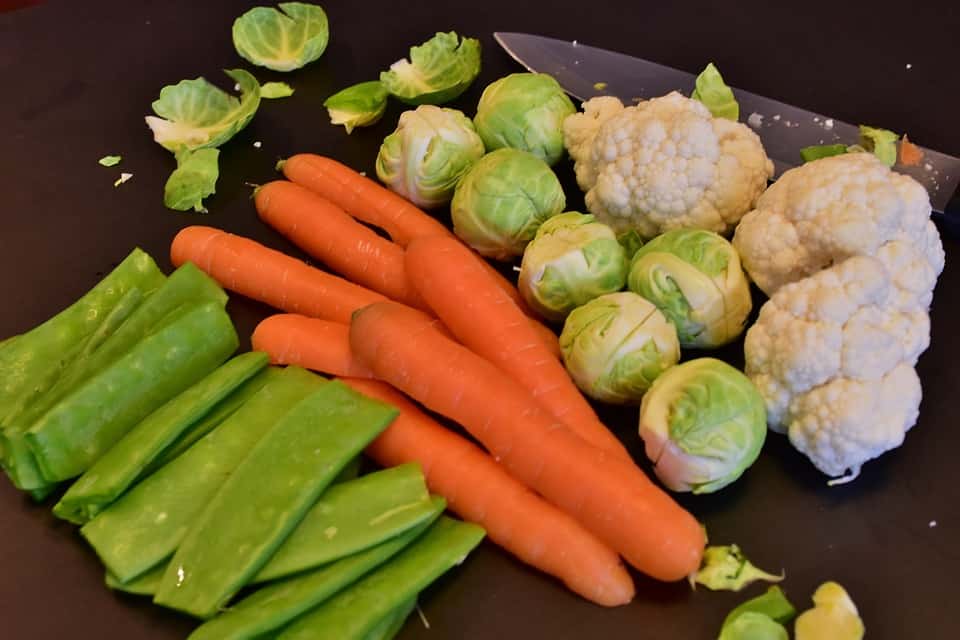 A selection of vegetables on a cutting board, including carrots, Brussels sprouts, cauliflower, and snap peas, with a knife beside them.