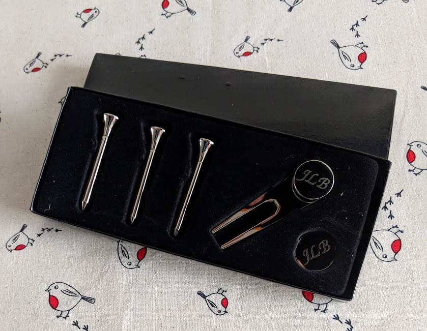 A black box containing three silver golf tees and a silver divot tool engraved with “jlb” on a white cloth with a bird pattern.