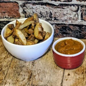 A white bowl of potato wedges next to a small red bowl filled with orange dipping sauce on a wooden surface in front of a brick wall.