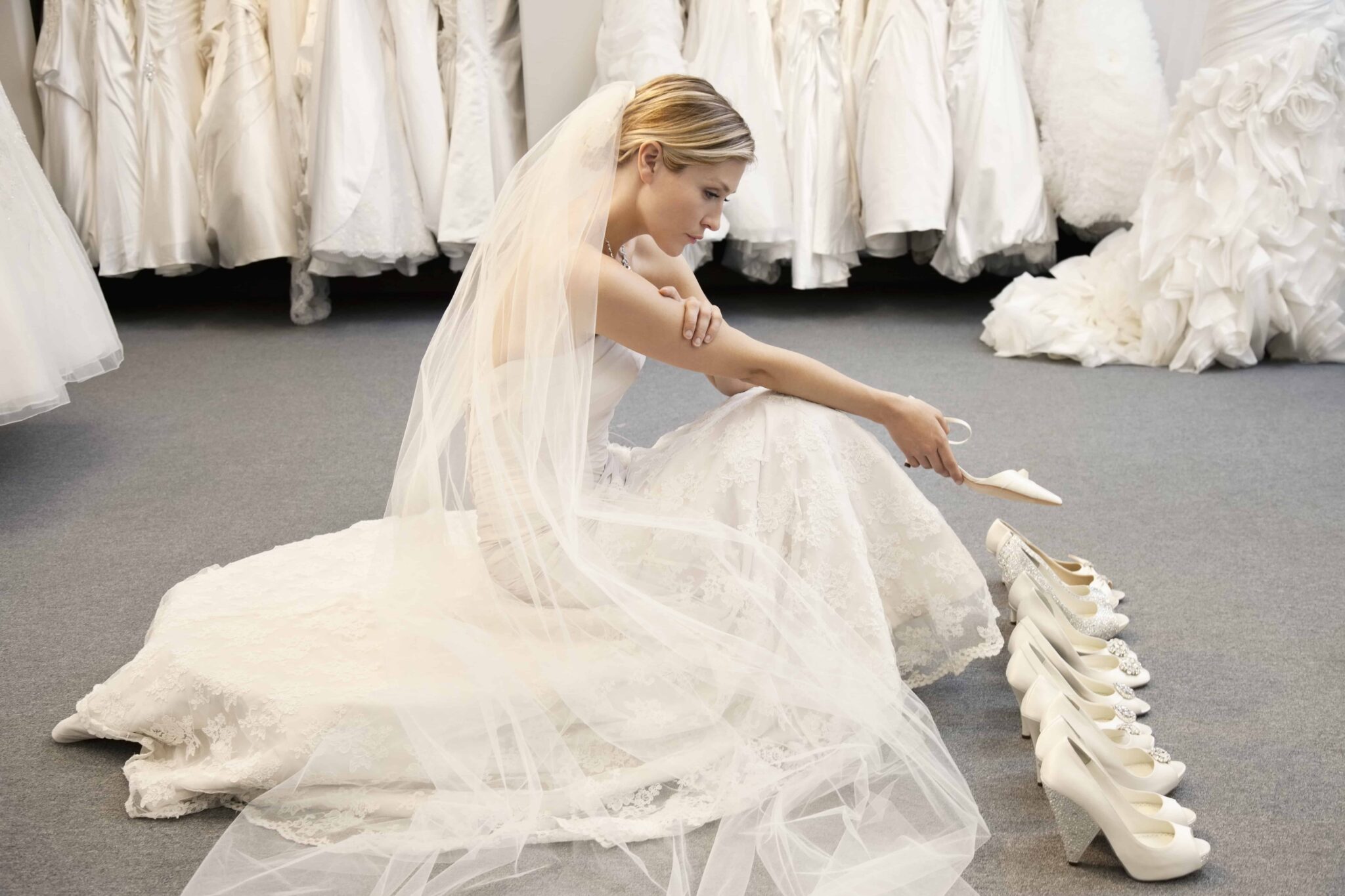 A bride in a stunning wedding dress sits elegantly on the floor, carefully adjusting her white shoes, surrounded by racks of exquisite wedding gowns.