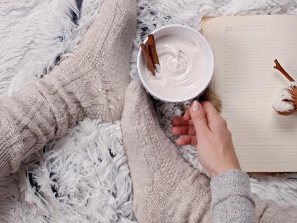 A sense of hygge fills the room as a person in cozy socks holds a cup of whipped coffee adorned with cinnamon sticks, next to an open notebook resting on a fuzzy white rug.