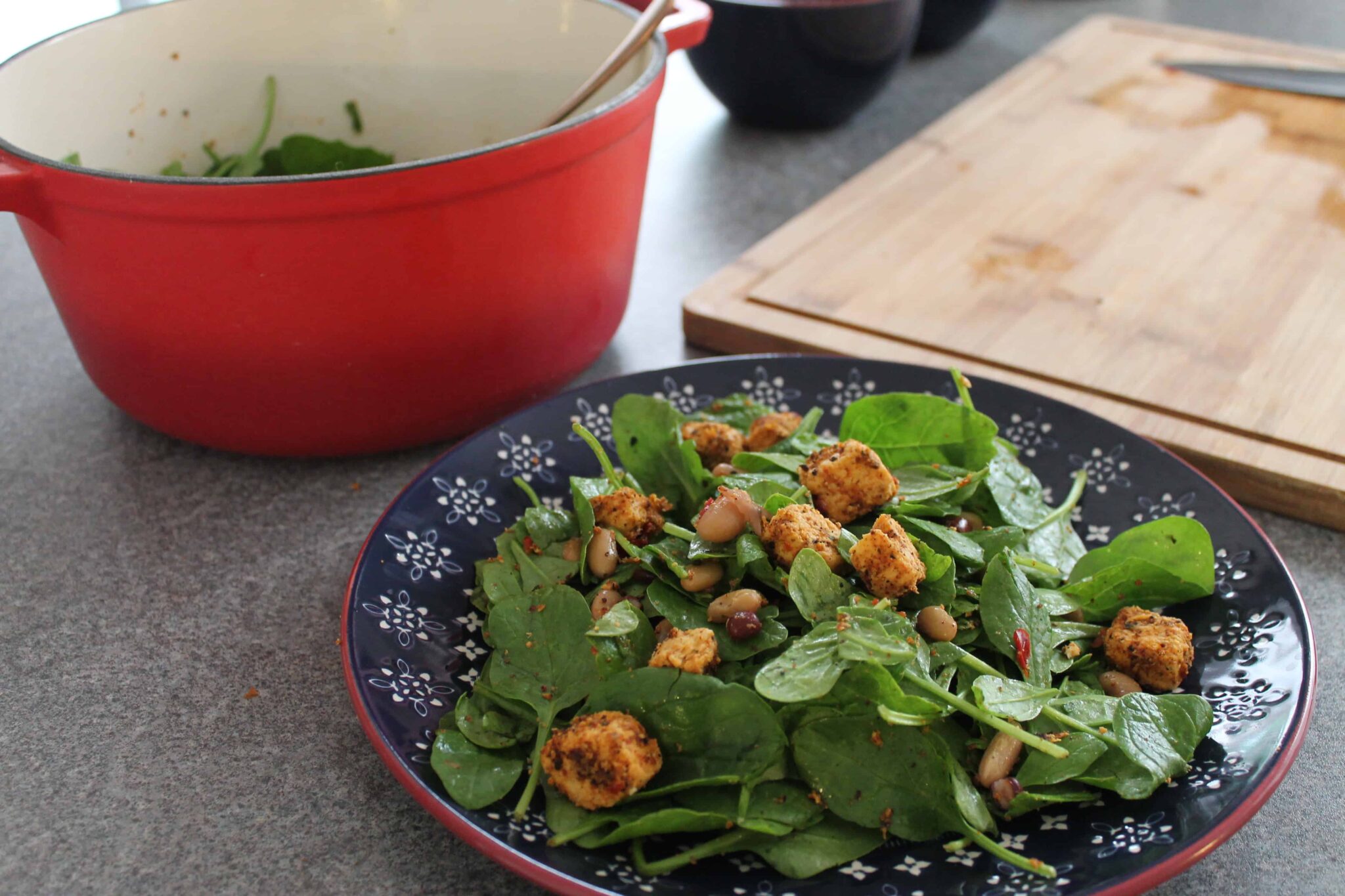 A plate of spinach salad with beans and croutons sits on a worktop, next to a red saucepan and a wooden chopping board.
