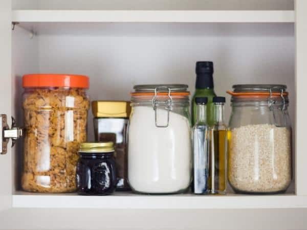 Inside of a food cupboard showing rice in a jar, pasta in a jar and flour in a jar with bottles of oil and jars of jam and coffee