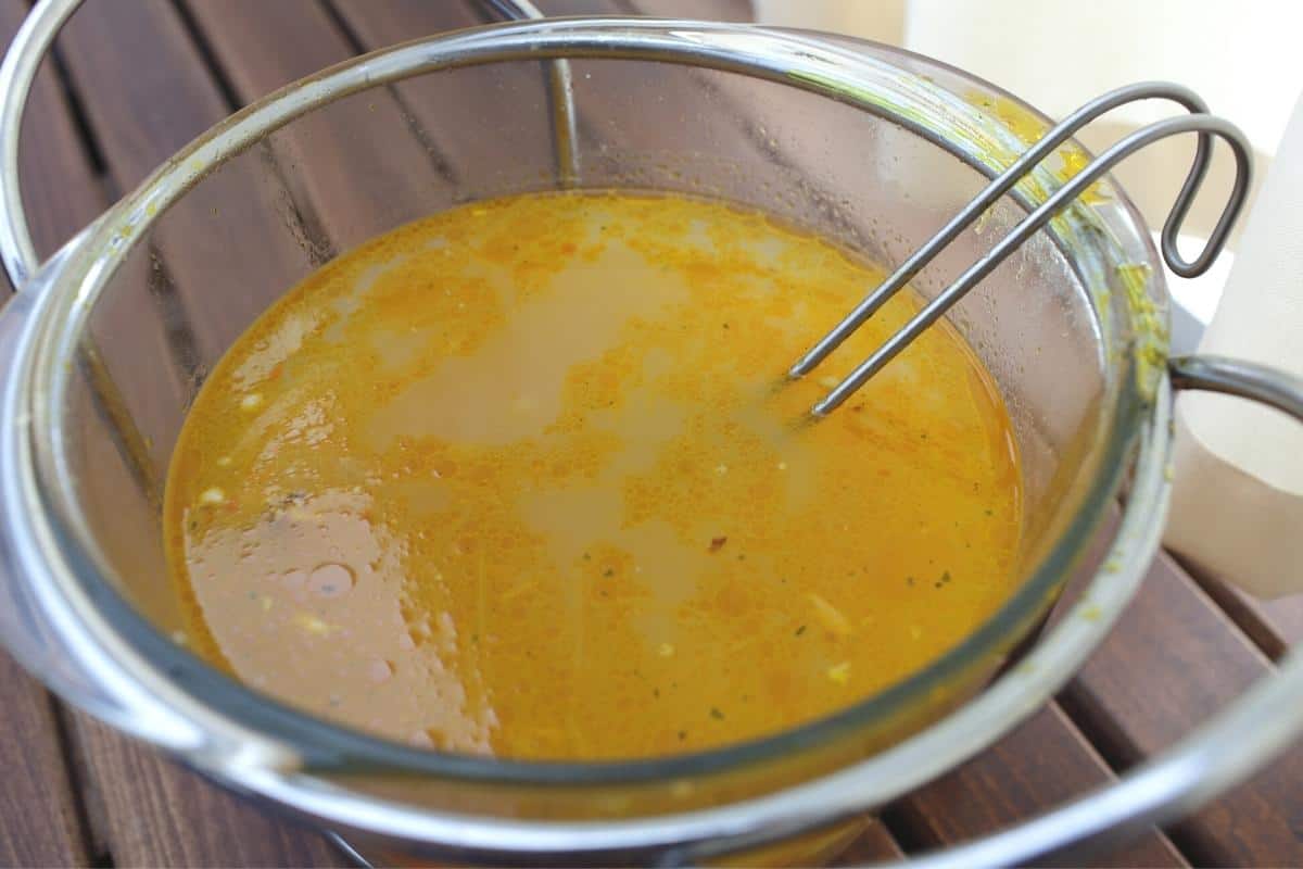 A glass bowl of vegetable stock with a metal whisk in.