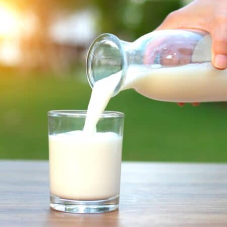 A hand pours milk from a glass jug into a clear glass on a wooden table, with a blurred green outdoor background.