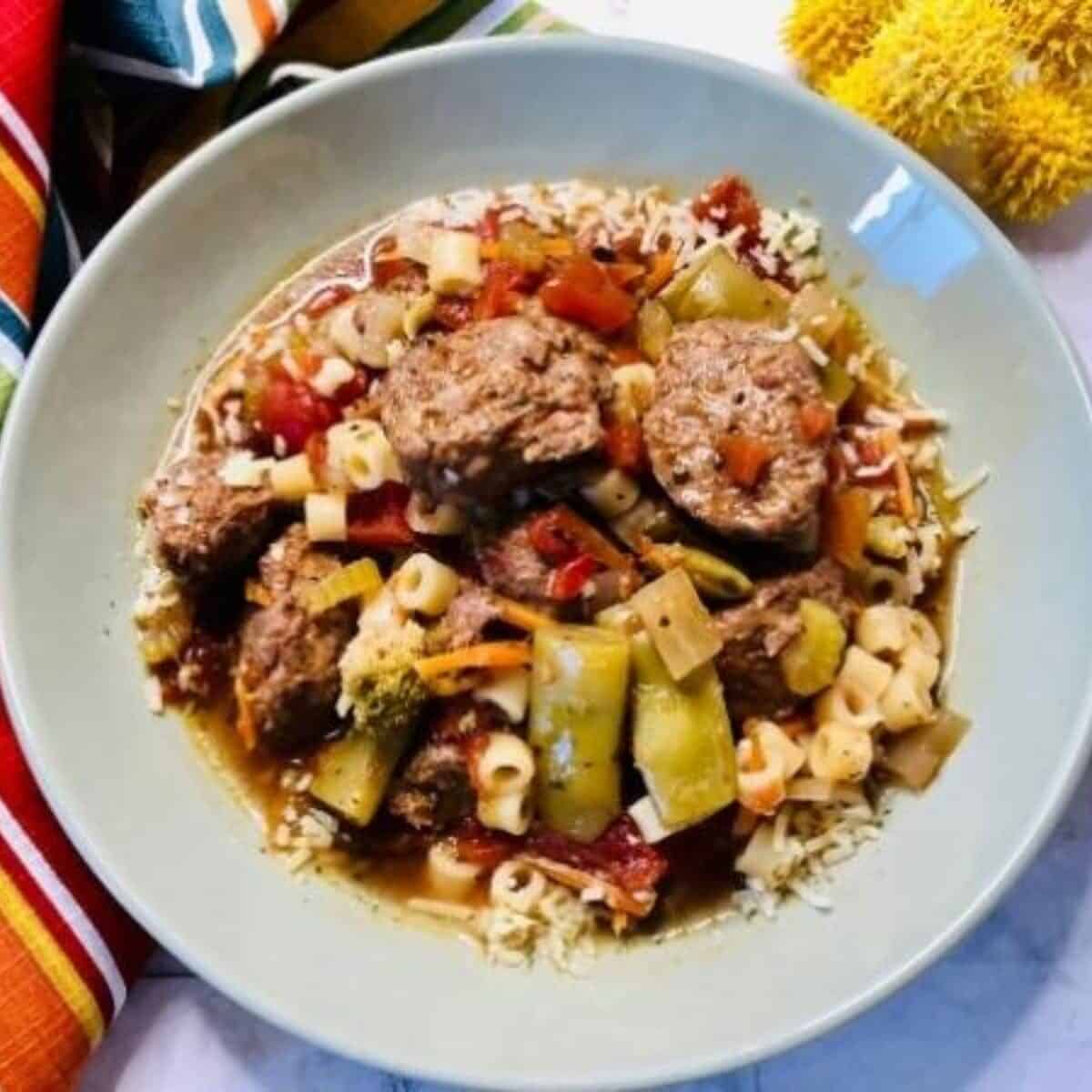 A bowl of soup with meatballs, pasta, diced tomatoes, vegetables, and grated cheese, served on a light green plate with a colourful striped serviette nearby.