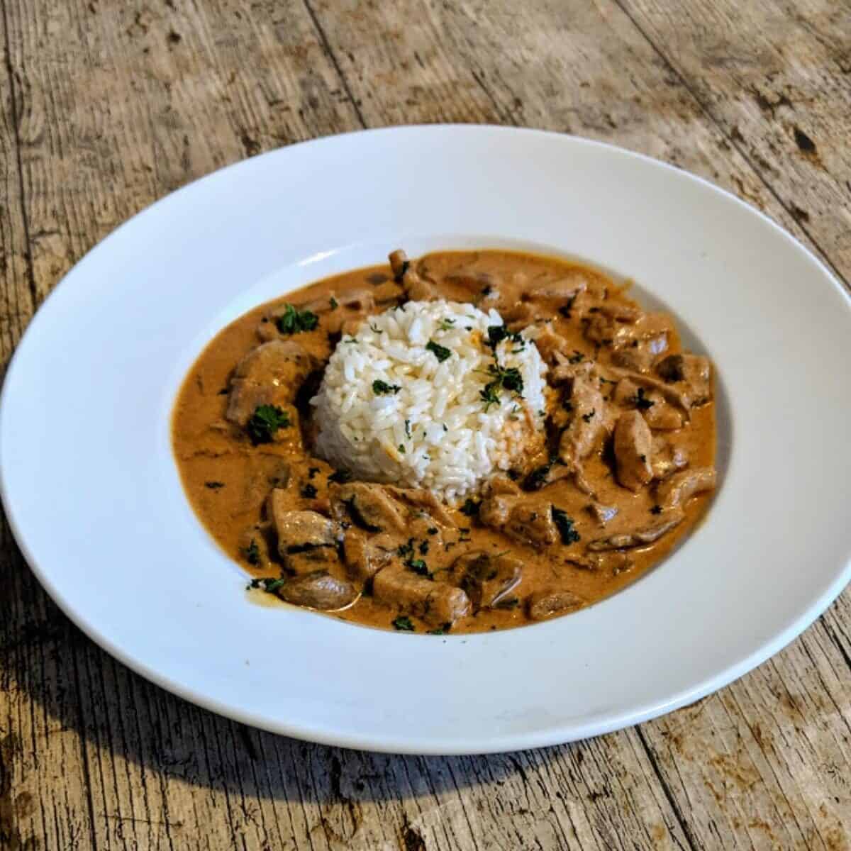 A white plate with a serving of rice in the centre, surrounded by a creamy brown stew with pieces of meat and mushrooms, garnished with chopped herbs, on a wooden table.