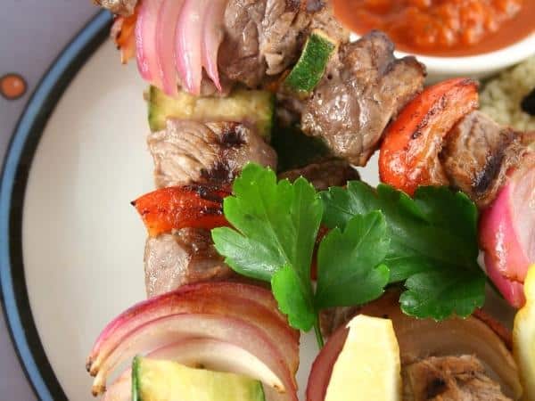 Close-up of grilled meat and vegetable skewers on a plate, garnished with parsley, with a sauce dish in the background.