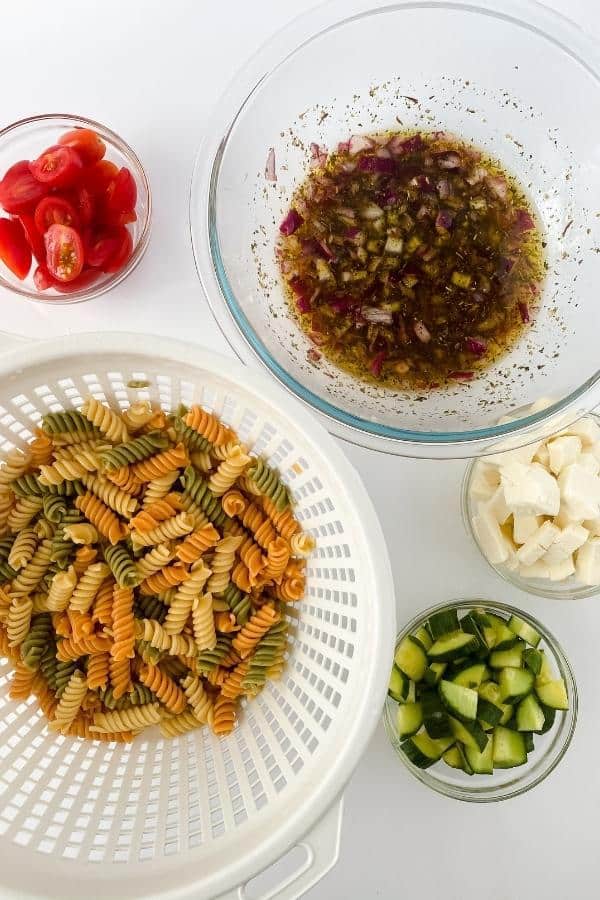 A bowl of salad dressing, bowls of sliced tomatoes, mozzarella, chopped cucumber, and a colander of tri-colour pasta spirals are arranged on a white surface.