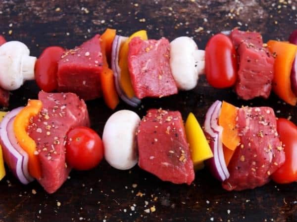 Raw beef skewers with red onions, bell peppers, cherry tomatoes, and mushrooms, seasoned with black pepper, on a dark surface.
