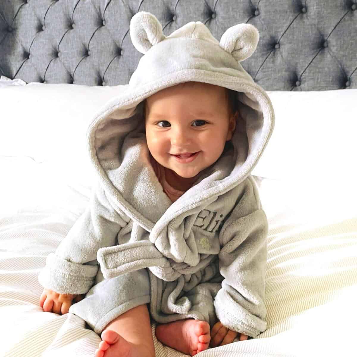 Baby sitting on a bed in a hooded dressing gown smiling at the camera.