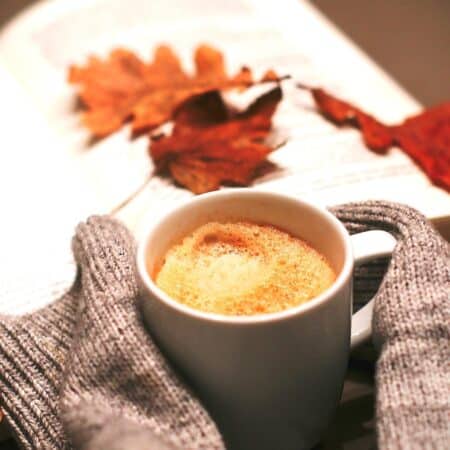 A white mug of coffee wrapped in grey knitted fabric sits in front of an open book with autumn leaves placed on its pages.