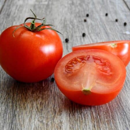 A whole tomato and a halved tomato sit on a wooden surface, with black peppercorns scattered in the background.