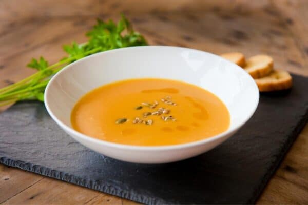 A white bowl of orange soup topped with seeds, placed on a black slate with parsley and sliced bread in the background.