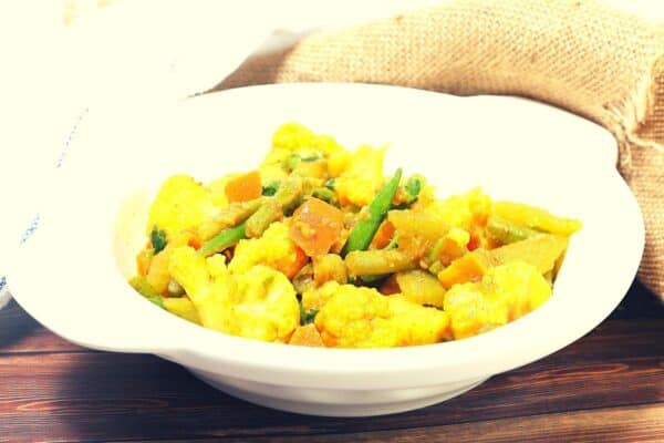 White bowl filled with a mixed vegetable curry, including cauliflower, carrots, green beans, and potatoes, placed on a wooden surface.