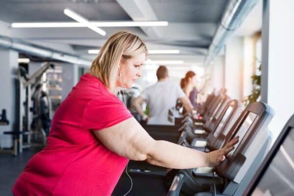 Woman in a pink shirt uses the touchscreen on a treadmill in a gym, with other people exercising in the background.