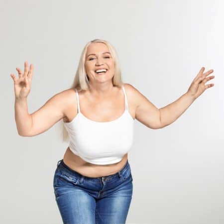 Smiling woman with long blonde hair in a white vest top and blue jeans stands with arms raised against a plain light background.
