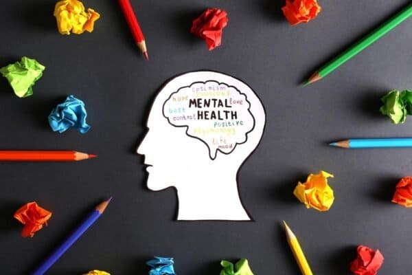 A paper cut-out of a head with mental health words inside is surrounded by coloured pencils and crumpled paper balls on a dark background.