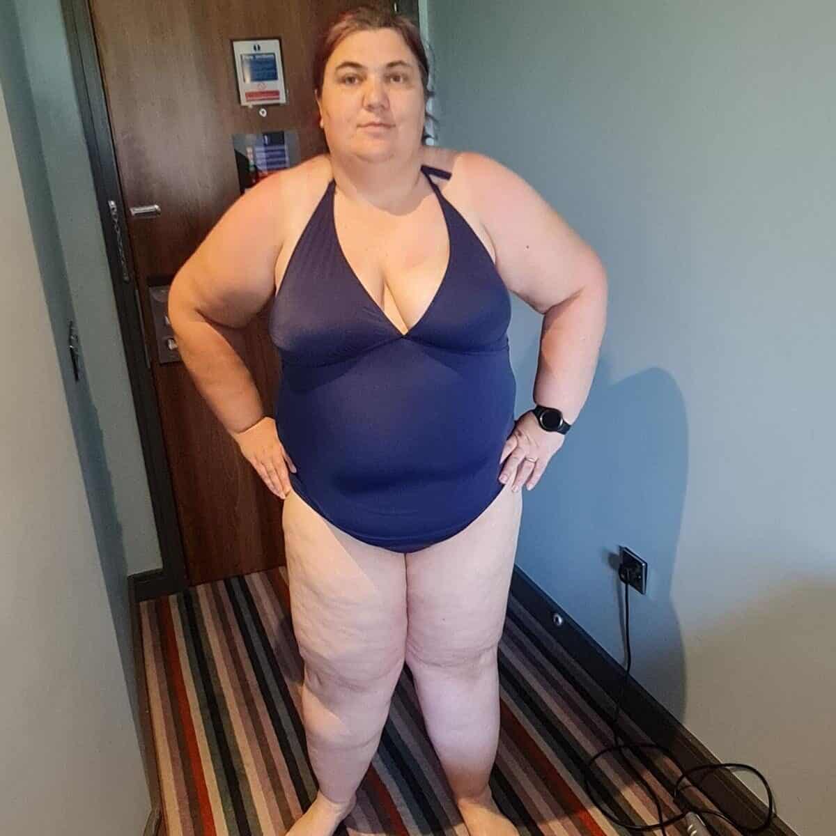 A person exudes body confidence in a blue swimsuit, standing on a striped carpet in the room, with hands confidently placed on hips.