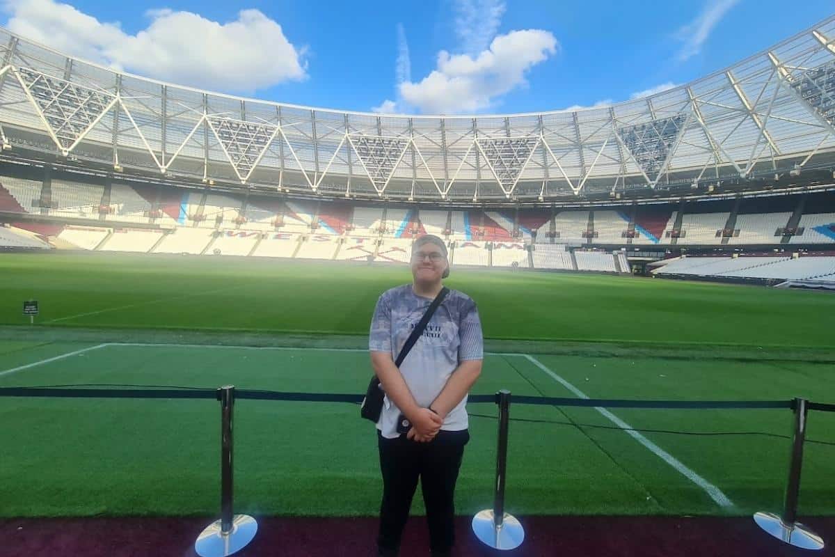 Ben, a teenage boy at the West Ham stadium tour with the pitch and seating behind him, he has a huge smile and is loving being there.