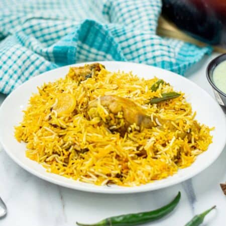 A plate of chicken biryani with pieces of chicken, basmati rice, green chillies, and spices, served on a white plate next to a blue checkered cloth.