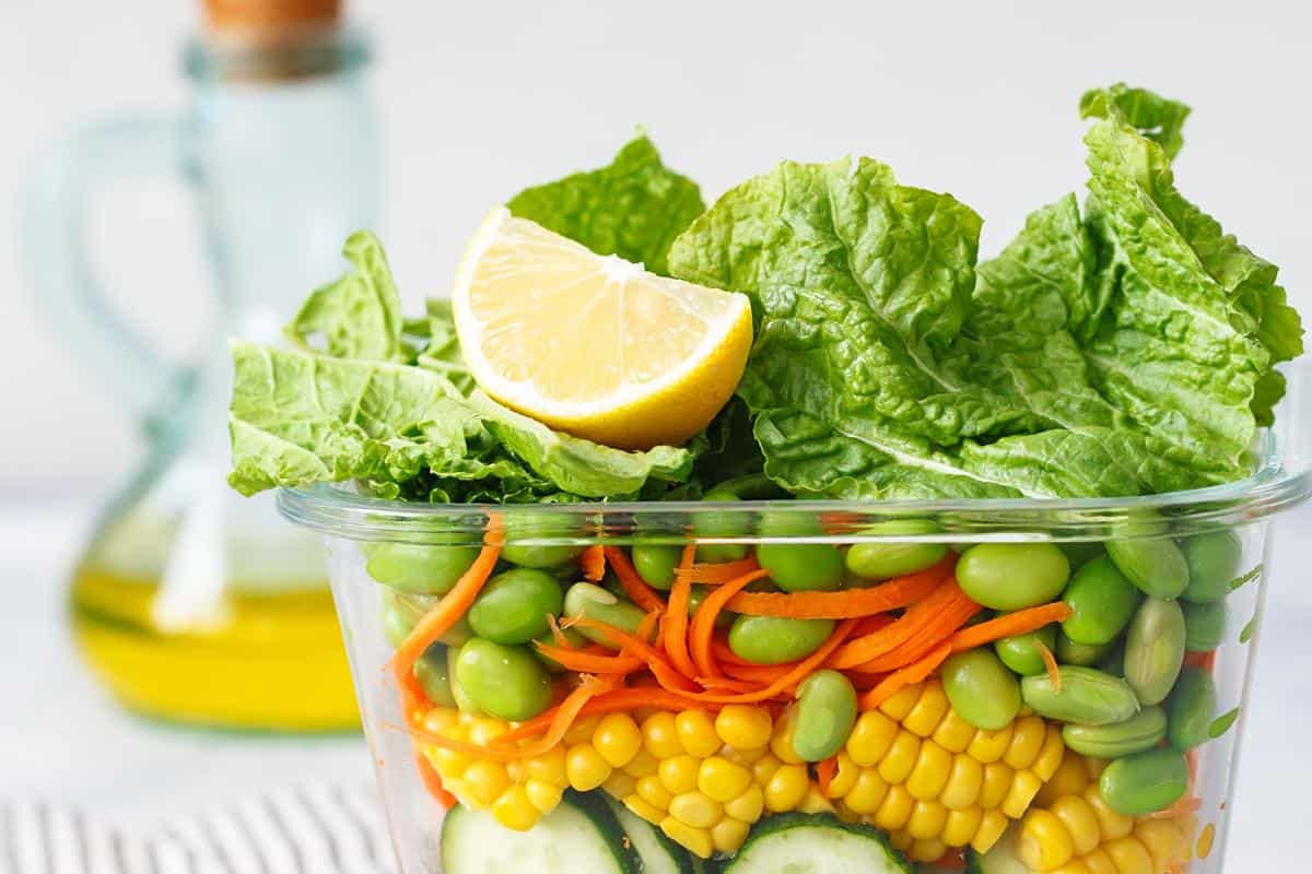 Plastic container of vegetables showing corn, grated carrot, cucumber, soy beans and lettuce topped with a chunk of lemon.