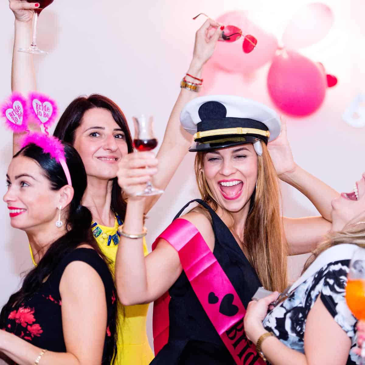 Ladies at a hen party wearing a pink sash, bride to be headband and with balloons holding wine glasses and looking drunk and as if they are having fun.