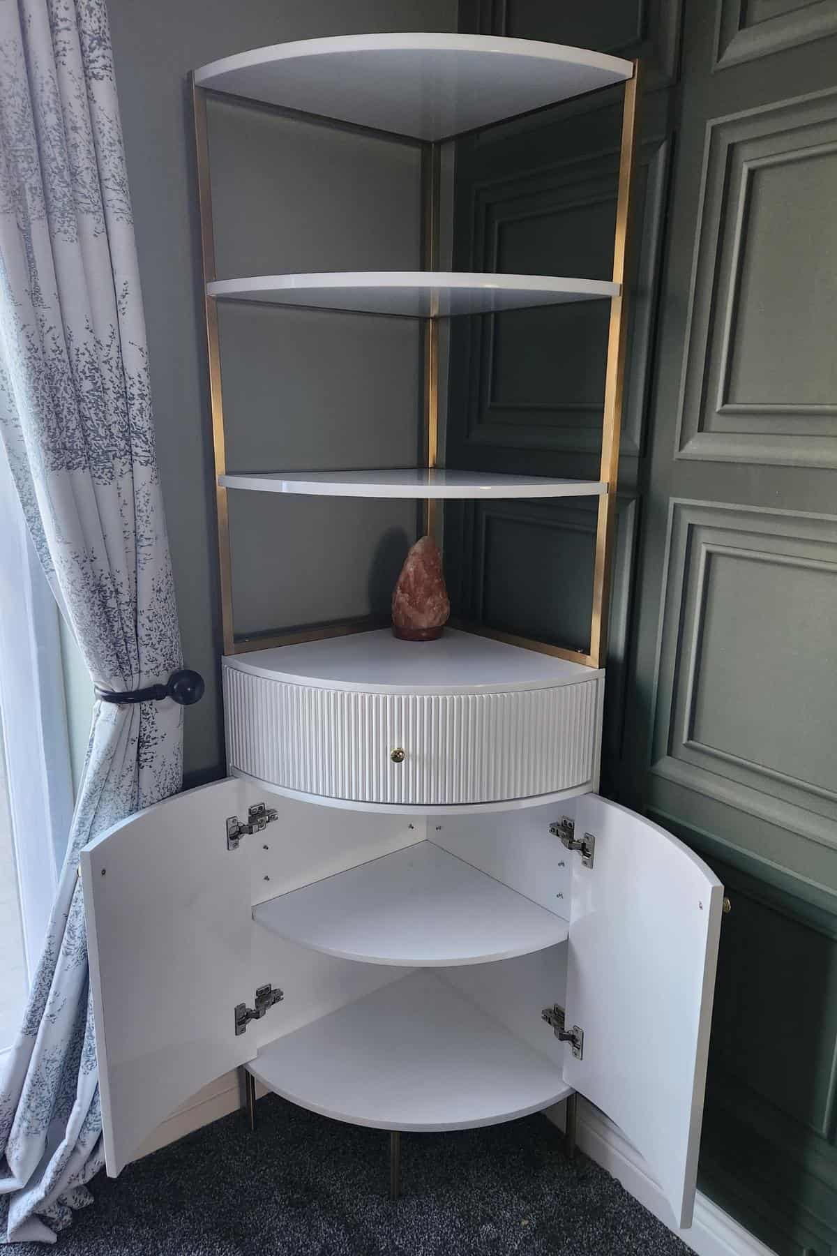 The corner shelving unit from Homary Furniture features elegant white shelves and a sleek gold frame. Open cabinet doors reveal additional white storage below, while a decorative stone graces one shelf, adding a touch of sophistication.