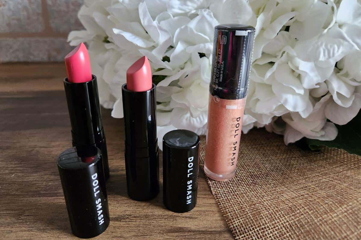 Two lipsticks and a lip gloss tube labeled "Doll Smash" are elegantly displayed on a wooden surface, with white hydrangeas adding a touch of charm in the background—perfect for your Valentine's Day look.