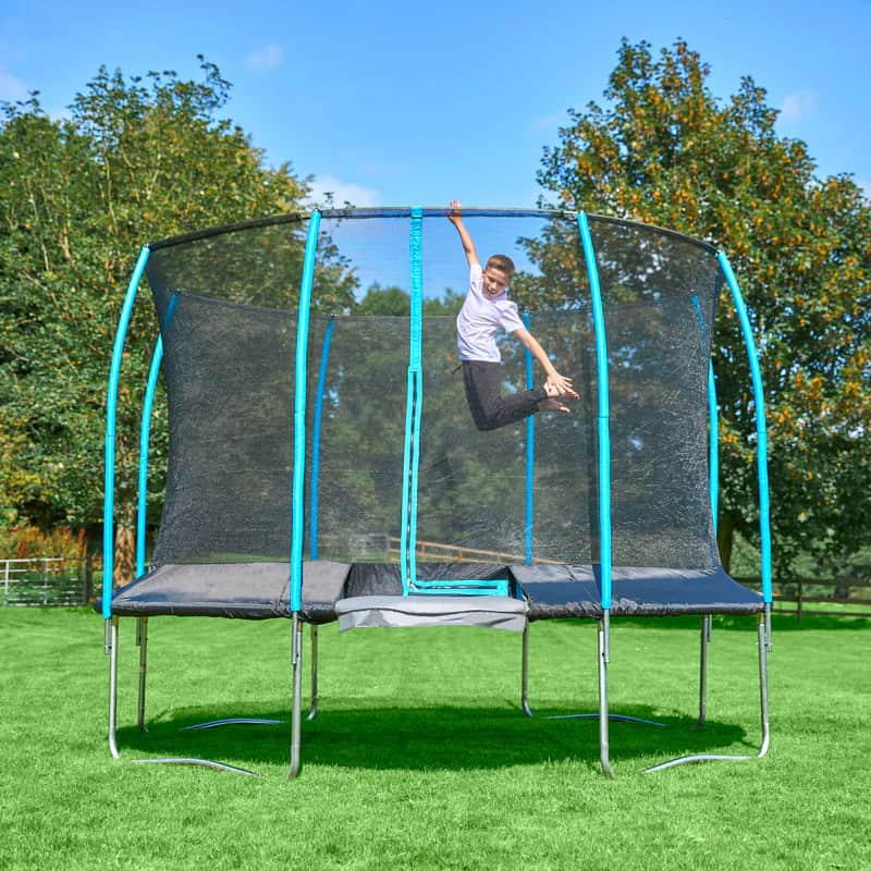 A child enjoys trampolining on a secure trampoline with a safety net, set in a grassy backyard. Towering trees frame the scene against a clear sky.