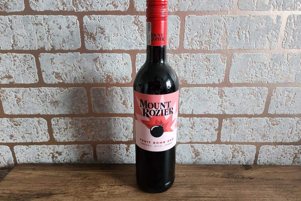 A bottle of Mount Rozier Fruit Bomb Red wine, perfect for Valentine's Day, with a red cap and label, placed on a wooden surface in front of a brick wall.
