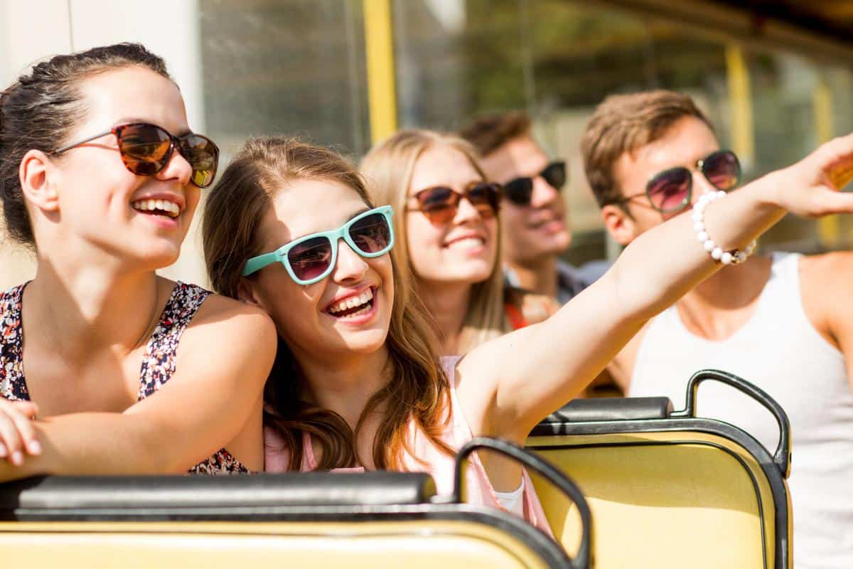 A group of young adults wearing sunglasses is on an open-top tour bus, soaking in the sunny day but staying safe in the sun with sunglasses.