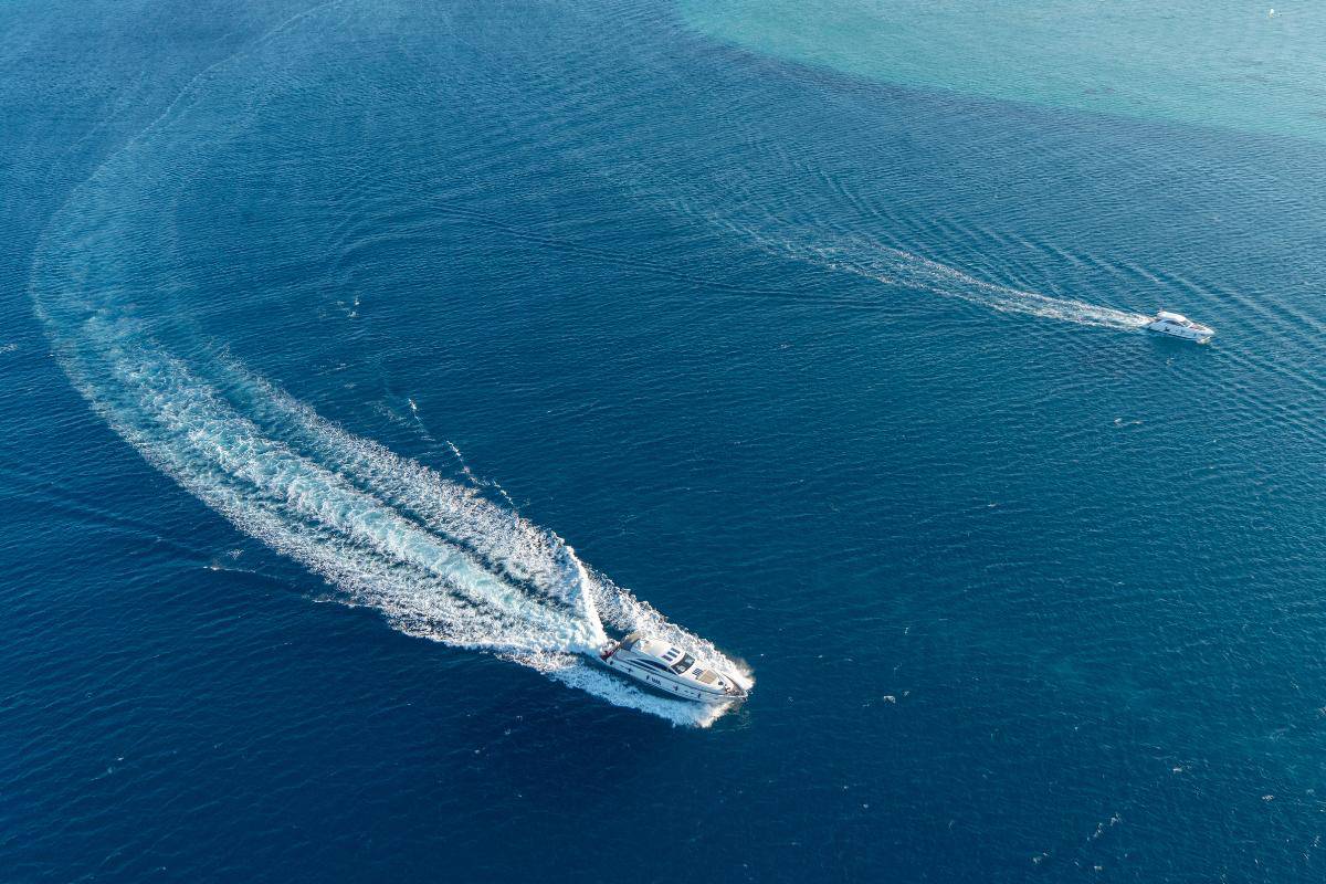 Two boats, including a sleek yacht, travel across the vast expanse of blue Mediterranean waters, leaving curved wakes behind them.