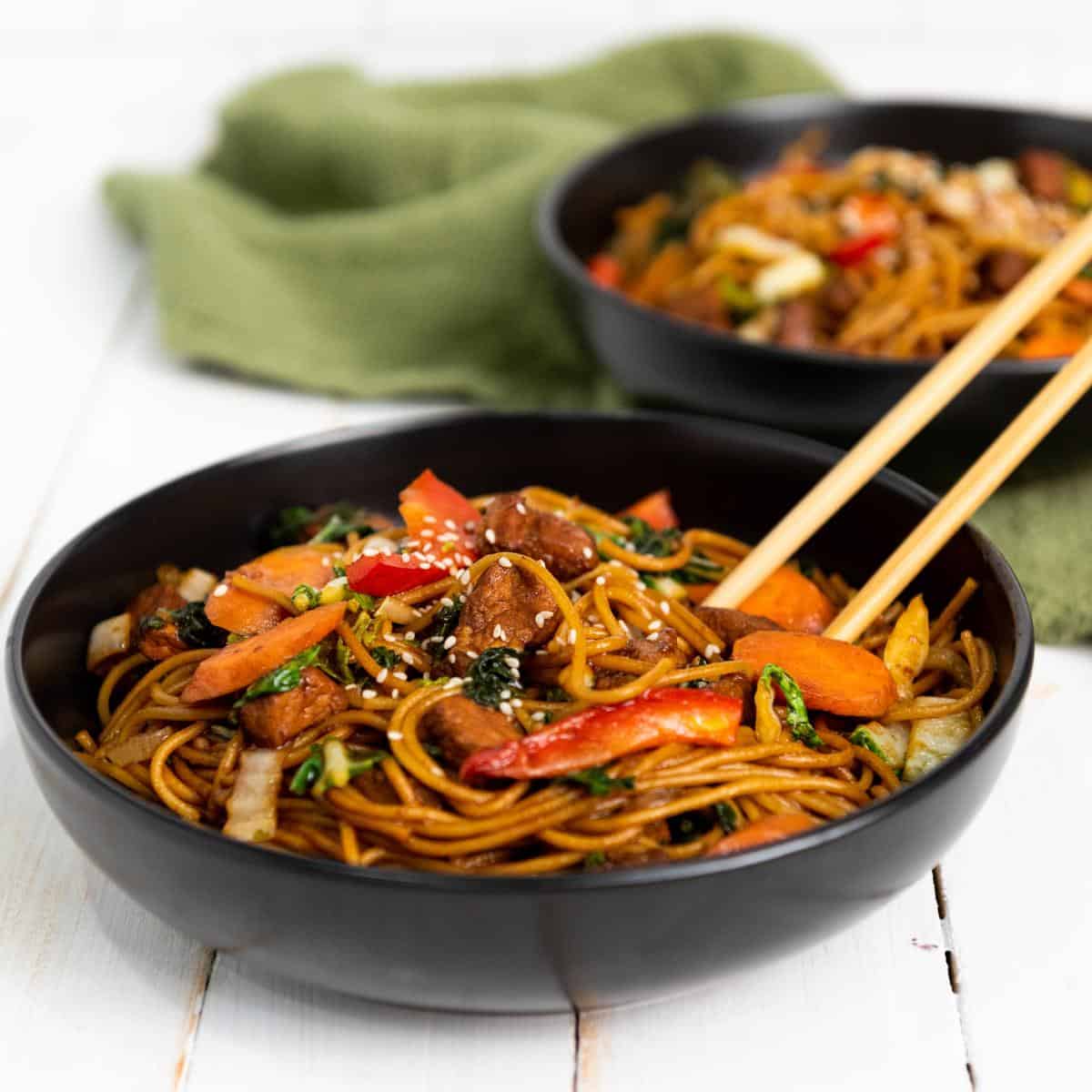 two bowls of pork lo mein in black bowls with chop sticks.