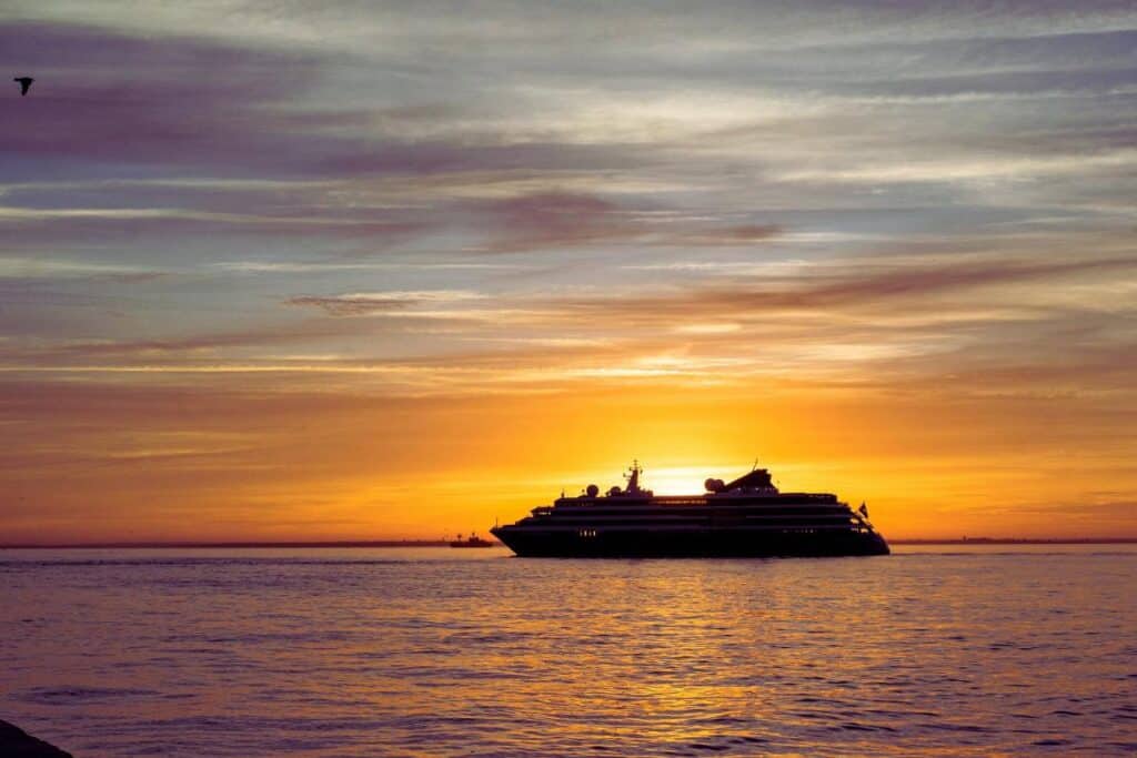 A large cruise ship sails on calm water at sunset, silhouetted against an orange and purple sky with scattered clouds—perfect inspiration for anyone planning a holiday cruise.