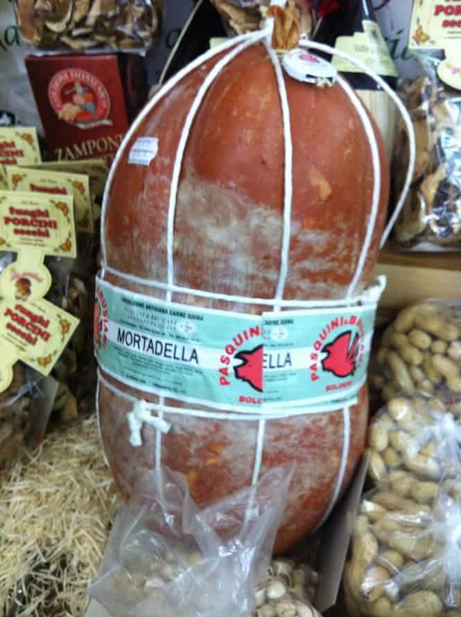 A large mortadella ham with white netting and label takes center stage on the display, surrounded by packages of nuts and mushrooms. This exquisite arrangement is reminiscent of a classic bologna feast.