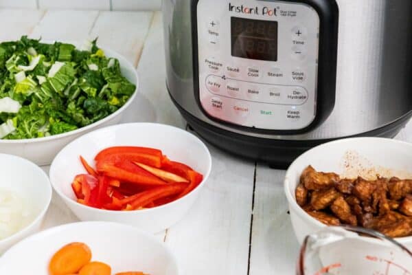 On the countertop sits an instant pot surrounded by bowls filled with sliced peppers, greens, onions, carrots, and tender cooked meat—perfect ingredients for a savory pork lo mein.