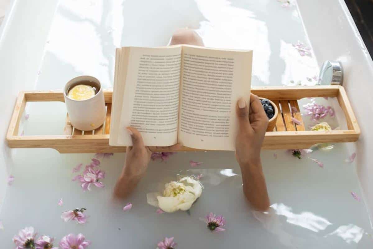 Enjoying a self care Sunday, a person relaxes in a bathtub, immersed in water dotted with floating flowers. A wooden tray nearby cradles a steaming mug and a bowl, while they indulge in reading their favourite book. A great one of the budget-friendly ways to treat yourself.