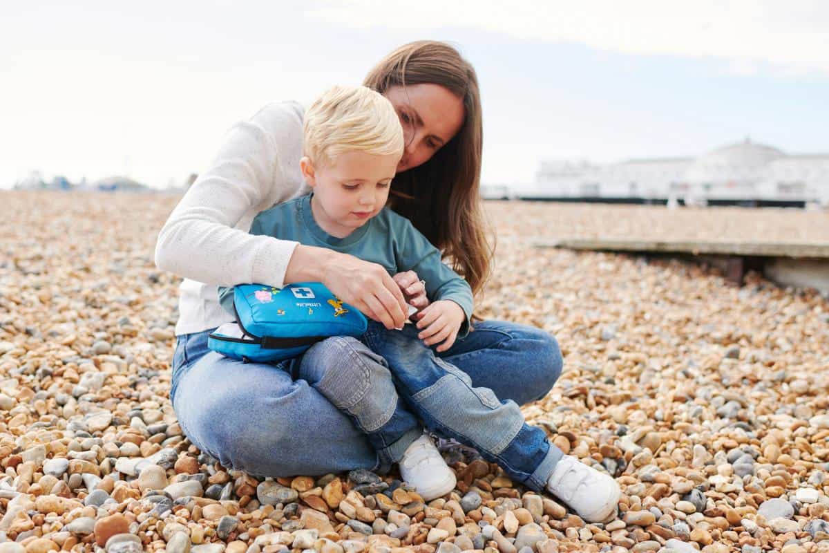 A woman and a child sit on a pebble-strewn beach, taking a break from their long car journey. The woman is helping the child with a blue backpack.