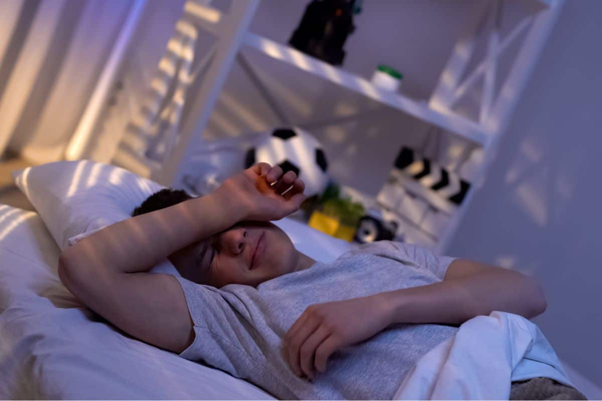 A smiling teenager in a gray shirt is lying on a bed, embodying the perfect blend of relaxation and teen bliss. His arm rests casually on his forehead, while a shelf with a soccer ball and clapperboard hints at his playful spirit.