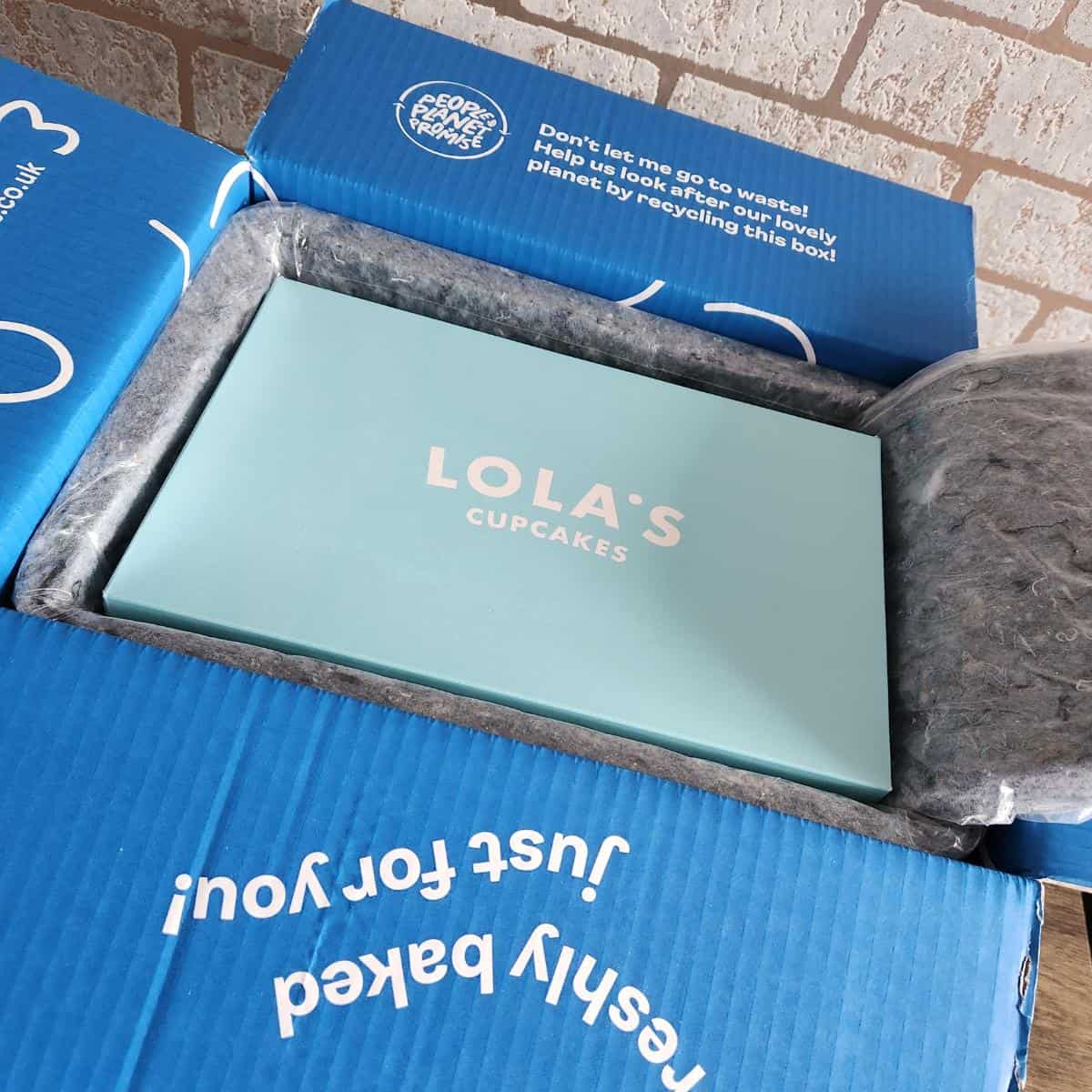 An open blue delivery box reveals a teal container labeled "Lola's Cupcakes," specifically designed for cupcakes by post. The box is insulated and lined with padding to ensure each delightful treat arrives in perfect condition.