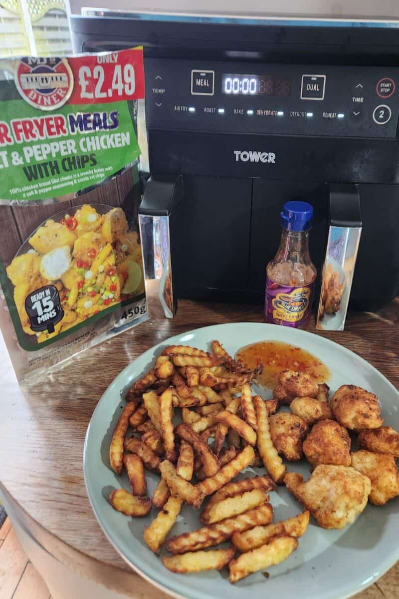 A plate of fries and chicken sits invitingly next to an air fryer, accompanied by a packet for salt and pepper chicken with chips, alongside a bottle of tangy hot-flavored sauce—perfect for quick and easy air fryer meals.