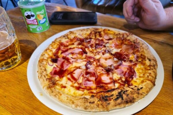 A pizza topped with cheese and meat, partially eaten, sits on a white plate. Nearby, on the wooden table often found in cozy hotels, are an open can of pringles, a smartphone, a hand reaching for more snacks, and a refreshing pint of beer.