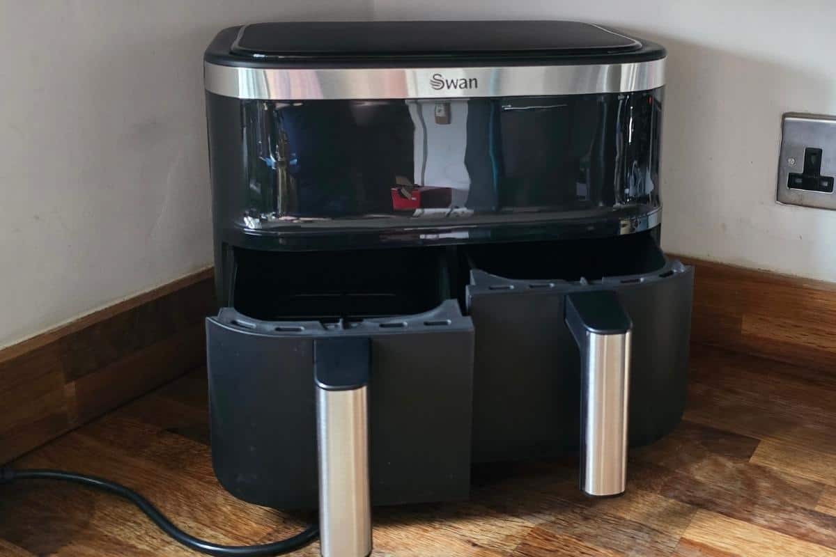 An air fryer with double basket sits on the wooden floor, its power cord plugged snugly into the wall socket—perfect as one of the university essentials for your teenager.