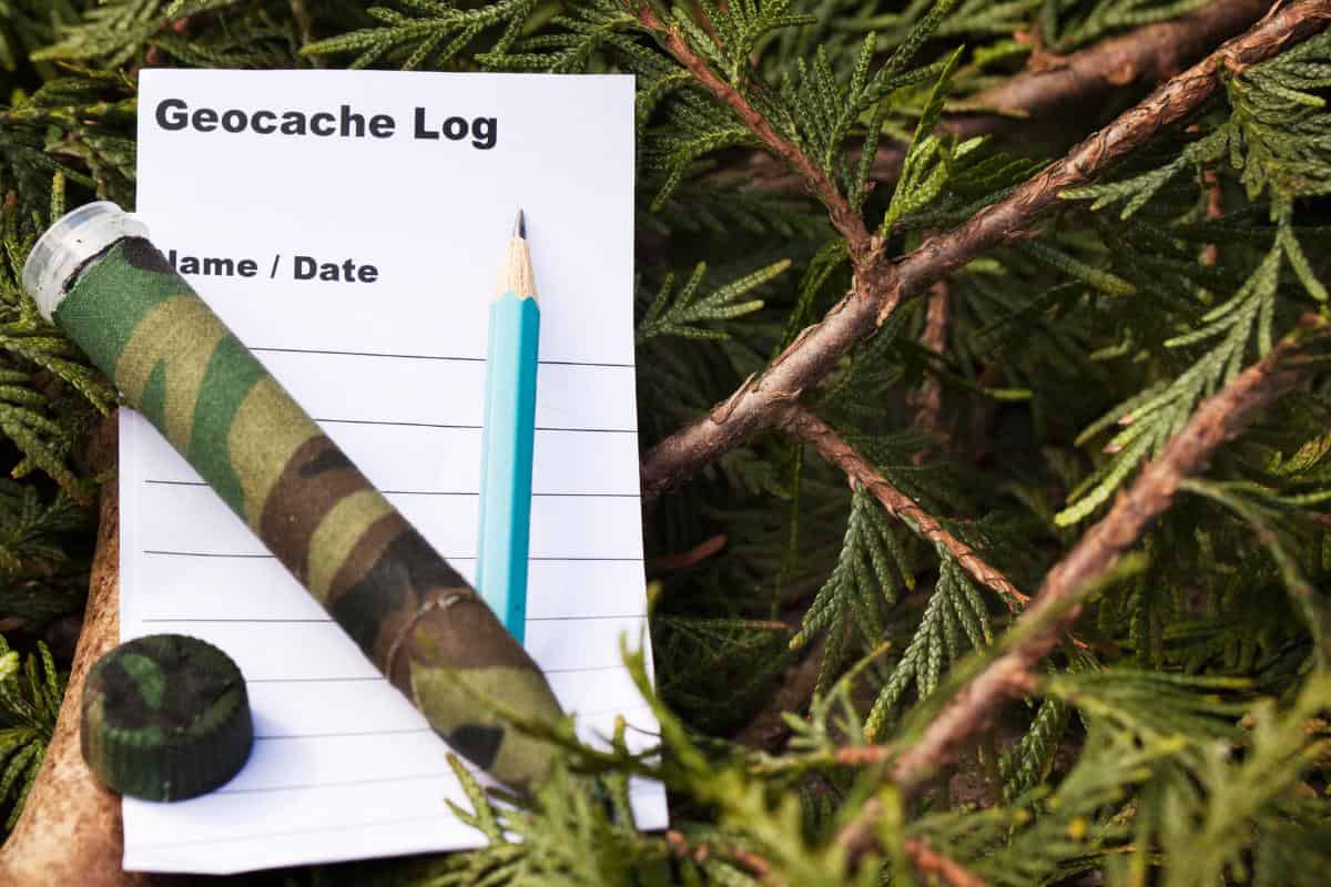 A tube covered in Camouflage fabric which has been opened to contain a pencil and a Geocache log with space to enter your name and date.