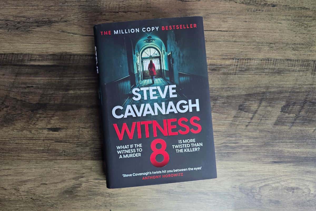 The book "Witness" by Steve Cavanagh rests on a wooden surface, its cover depicting a hallway with a figure in red. A captivating addition to any collection, this gripping novel invites readers into a world of suspense, reflecting the power books have to transport and thrill.