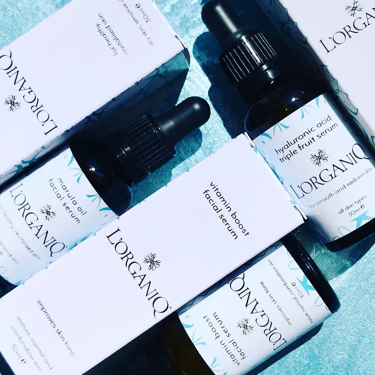 L'Organico skincare bottles and boxes elegantly arranged on a blue surface, showcasing "vitamin boost facial serum" and "hyaluronic acid triple fruit serum." This refined Serum Trio collection by L'ORGANIQ is available for just £80, promising to elevate your skincare routine.