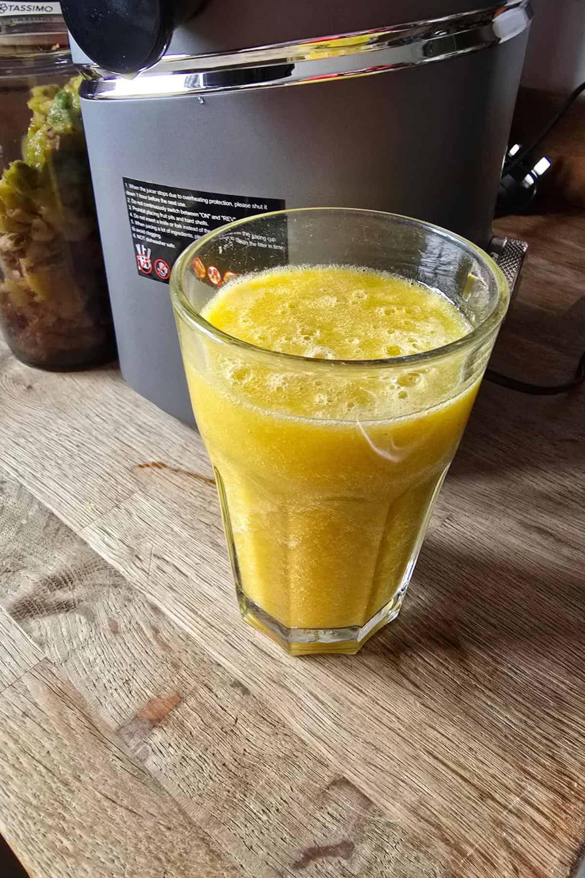 A clear glass filled with frothy yellow juice sits on a wooden countertop. Behind it, a metal juicer and a jar containing vegetable scraps are partially visible, highlighting that the juice was freshly made with the juicer.