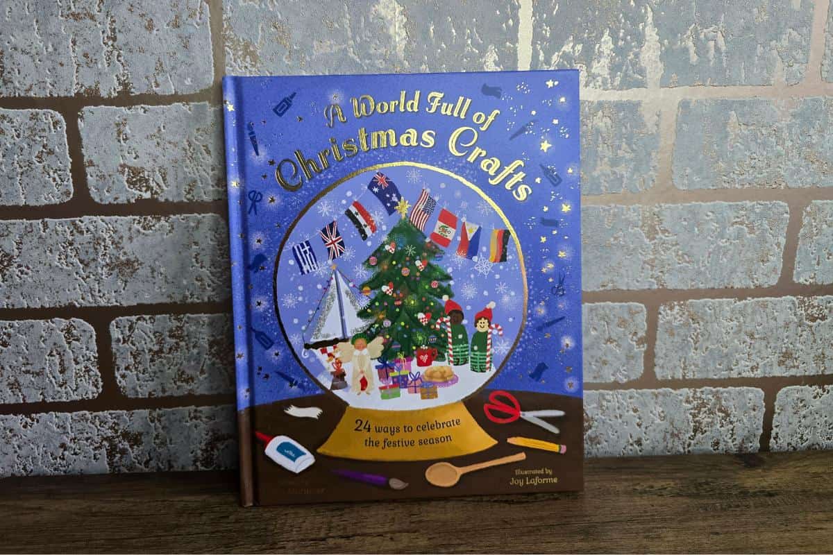 Cover of a charming book titled "A World Full of Christmas Crafts," ideal for children, featuring illustrations of a Christmas tree, Santa, various flags, and crafting tools against a brick wall background.